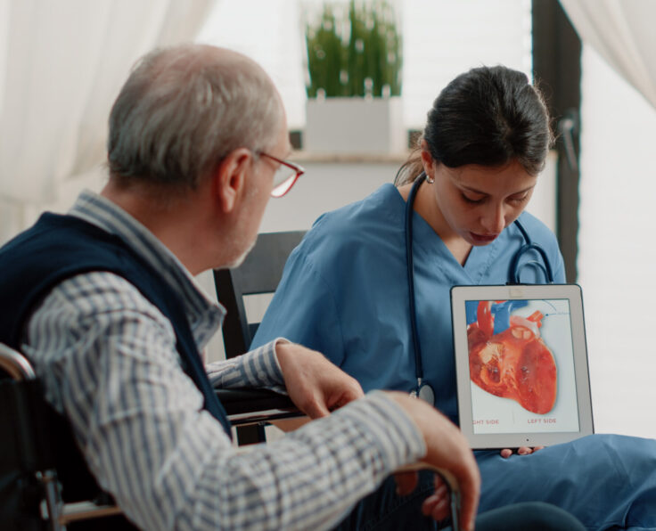 Nurse holding tablet with heart figure for cardiology diagnosis, showing cardiovascular issues to disabled patient at facility. Retired man sitting in wheelchair looking at blood vessel