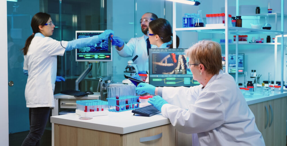 Nurse holding tablet computer with scientific informations while chemist using microscope with chemical test tube near, in lab background at night. Medical science research and development concept